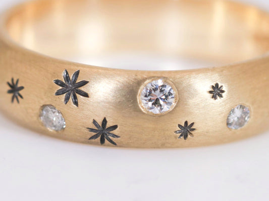 A rounded yellow gold ring band with hand carved, black stars sprinkled between 3 bright white full cut diamonds that are flush set into the gold.
