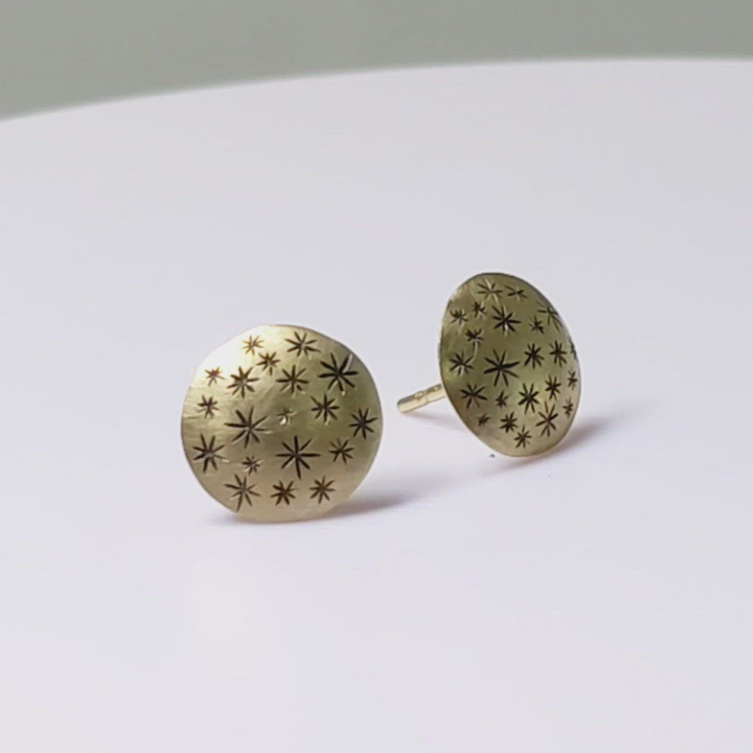 A video of a pair of 14k yellow gold earrings that are 2 domed circles adorned with hand carved black stars spinning on a turntable.