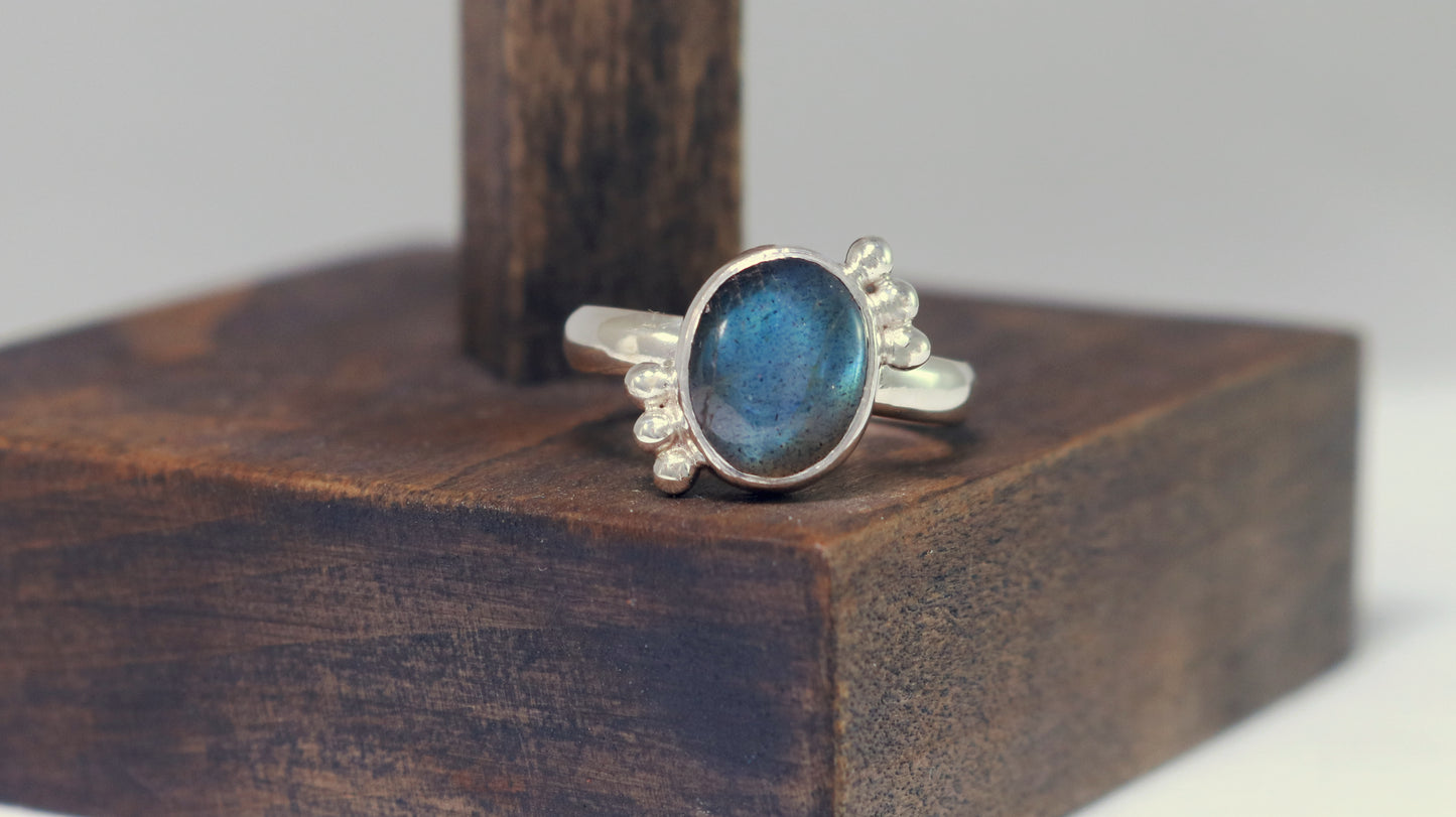 A royal blue, oval labradorite stone is bezel set on a sterling silver ring. On opposite diagonals are three silver bubbles with a total of 6 bubbles on the stone.