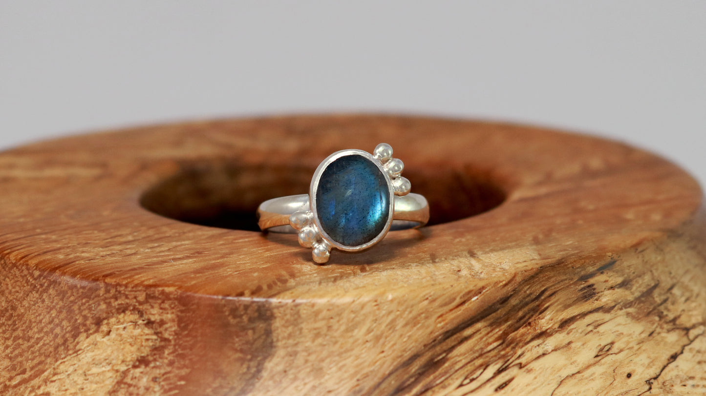 A royal blue, oval labradorite stone is bezel set on a sterling silver ring. On opposite diagonals are three silver bubbles with a total of 6 bubbles on the stone.