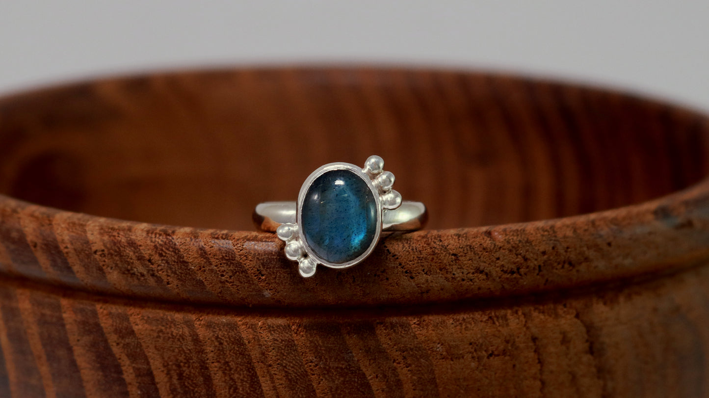 A royal blue, oval labradorite stone is bezel set on a sterling silver ring. On opposite diagonals are three silver bubbles with a total of 6 bubbles on the stone.