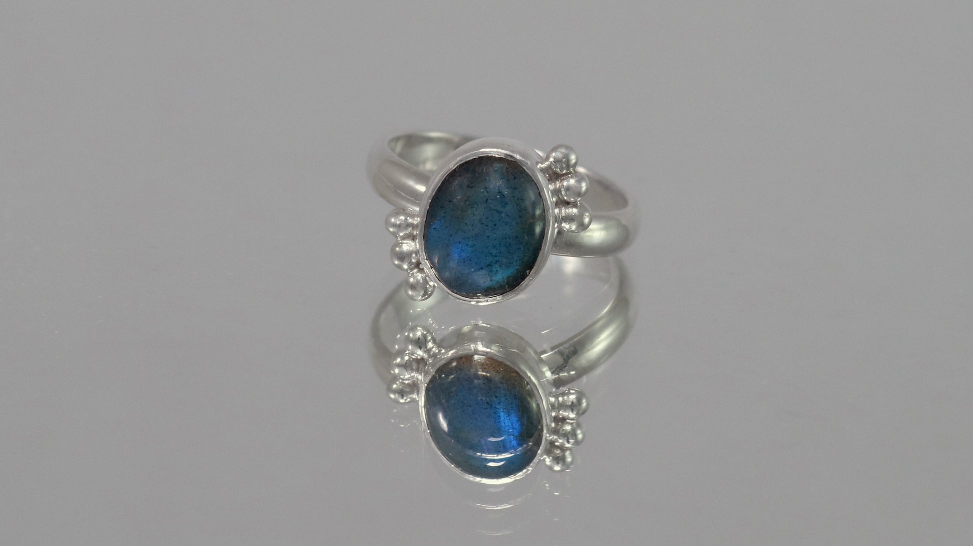 A royal blue, oval labradorite stone is bezel set on a sterling silver ring. On opposite diagonals are three silver bubbles with a total of 6 bubbles on the stone.
