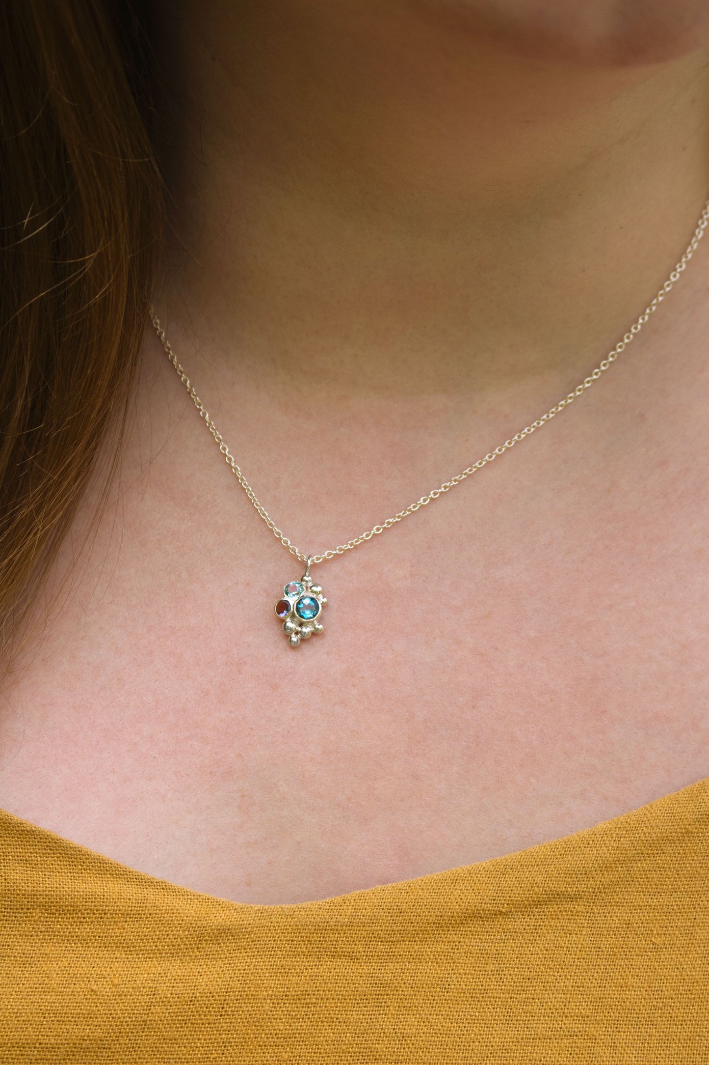 A model wearing a pendant on a silver chain, three round gemstones, all a different shade of blue, set in a pyramid shape with randomly placed silver bubbles.