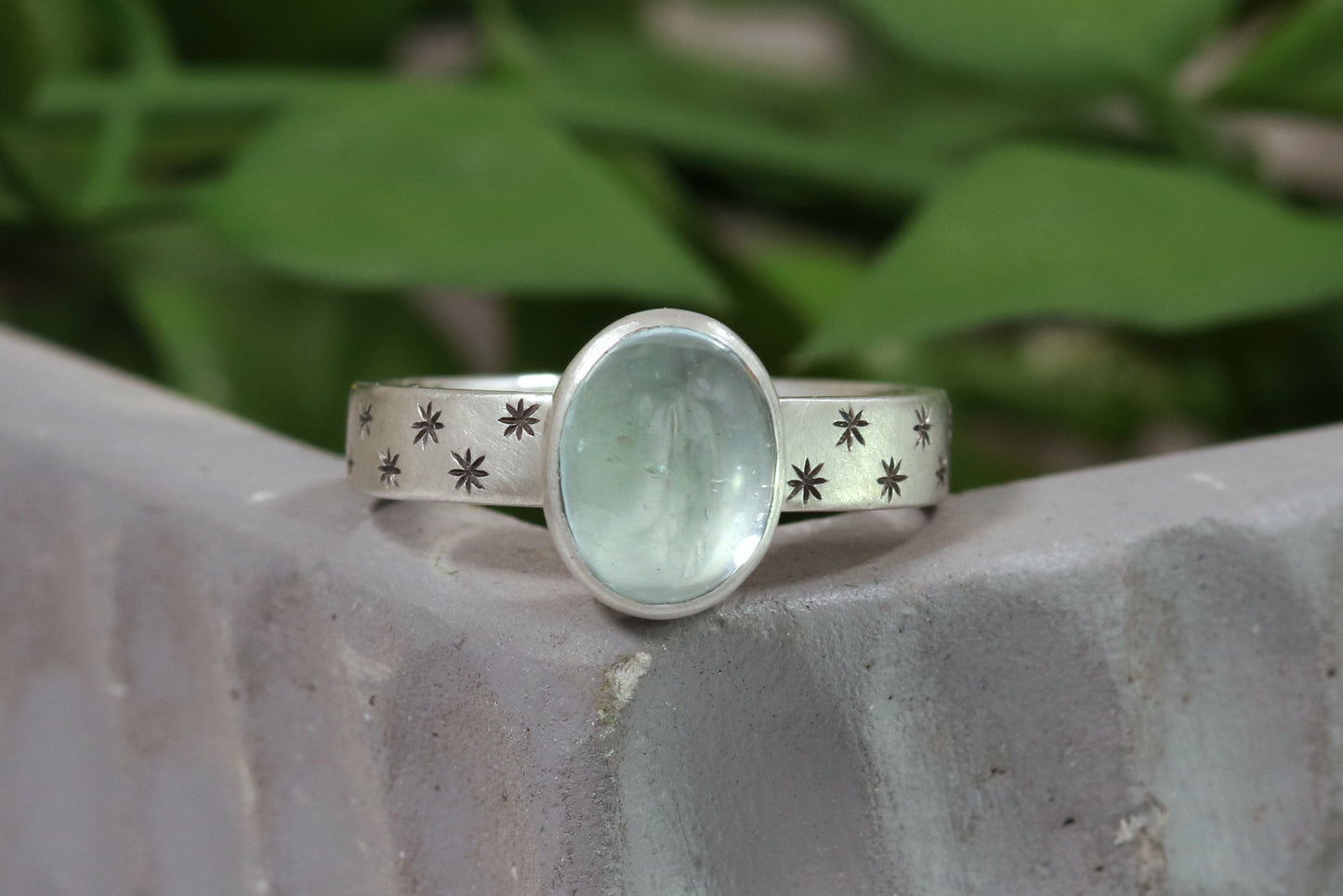 Oval natural gemstone set in sterling silver bezel on a wide sterling silver ring band with checker board of hand cut black stars on either side.