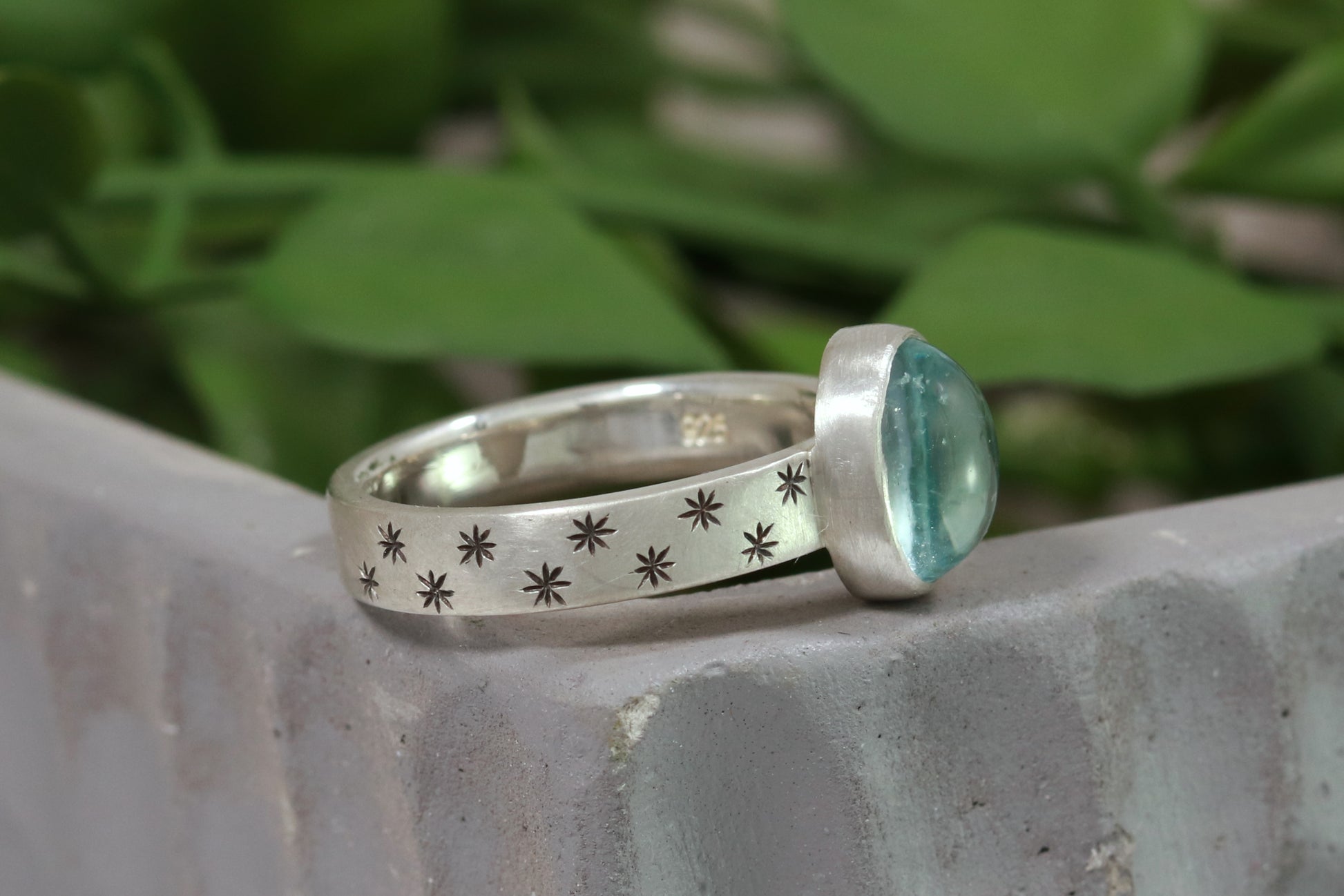 Oval natural gemstone set in sterling silver bezel on a wide sterling silver ring band with checker board of hand cut black stars on either side.