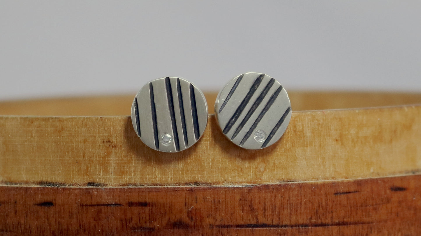 Round silver disc studs with black lines randomly spaced and a single white diamond flush set at the edge.