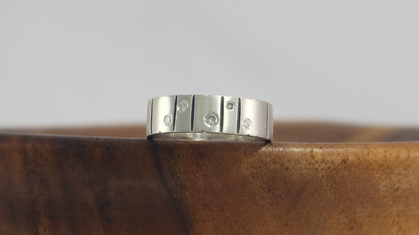 A thick silver ring band with randomly spaced black line details is flush set with 5 various sized diamonds.
