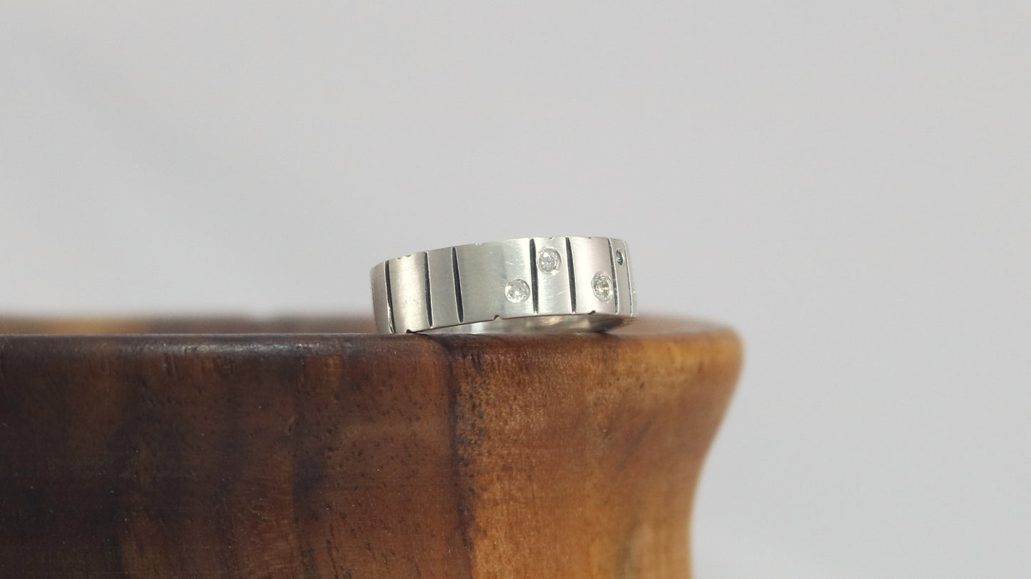 A thick silver ring band with randomly spaced black line details is flush set with 5 various sized diamonds.