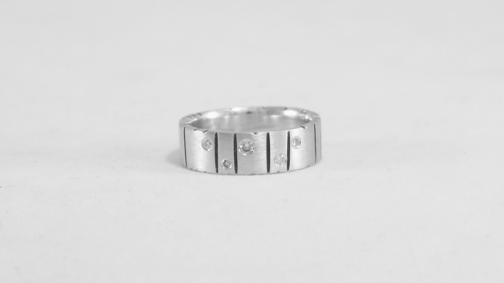 A thick silver ring band with randomly spaced black line details is flush set with 5 various sized diamonds.