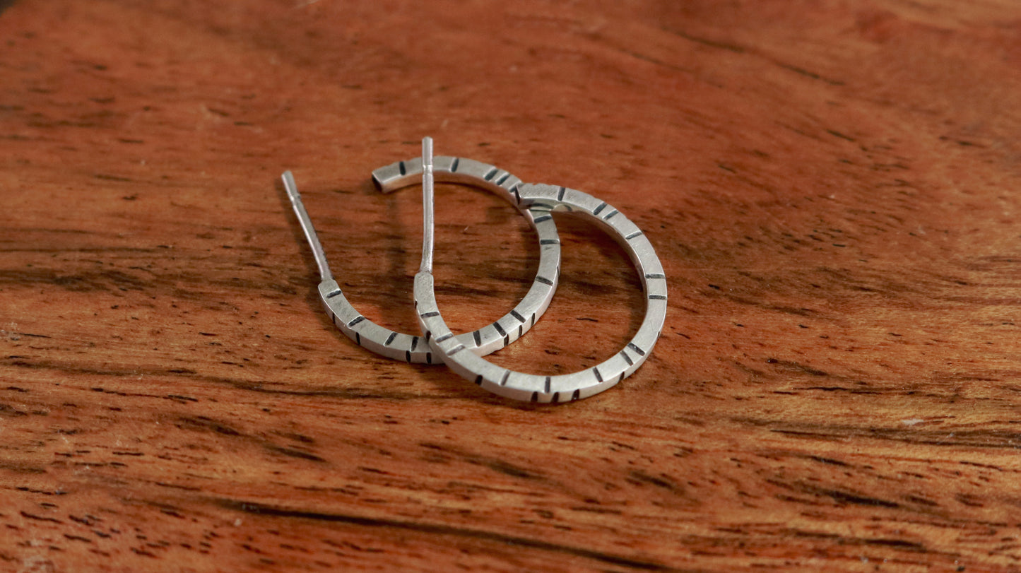 Thin sterling silver hoop earrings with randomly spaced black lines on each side.