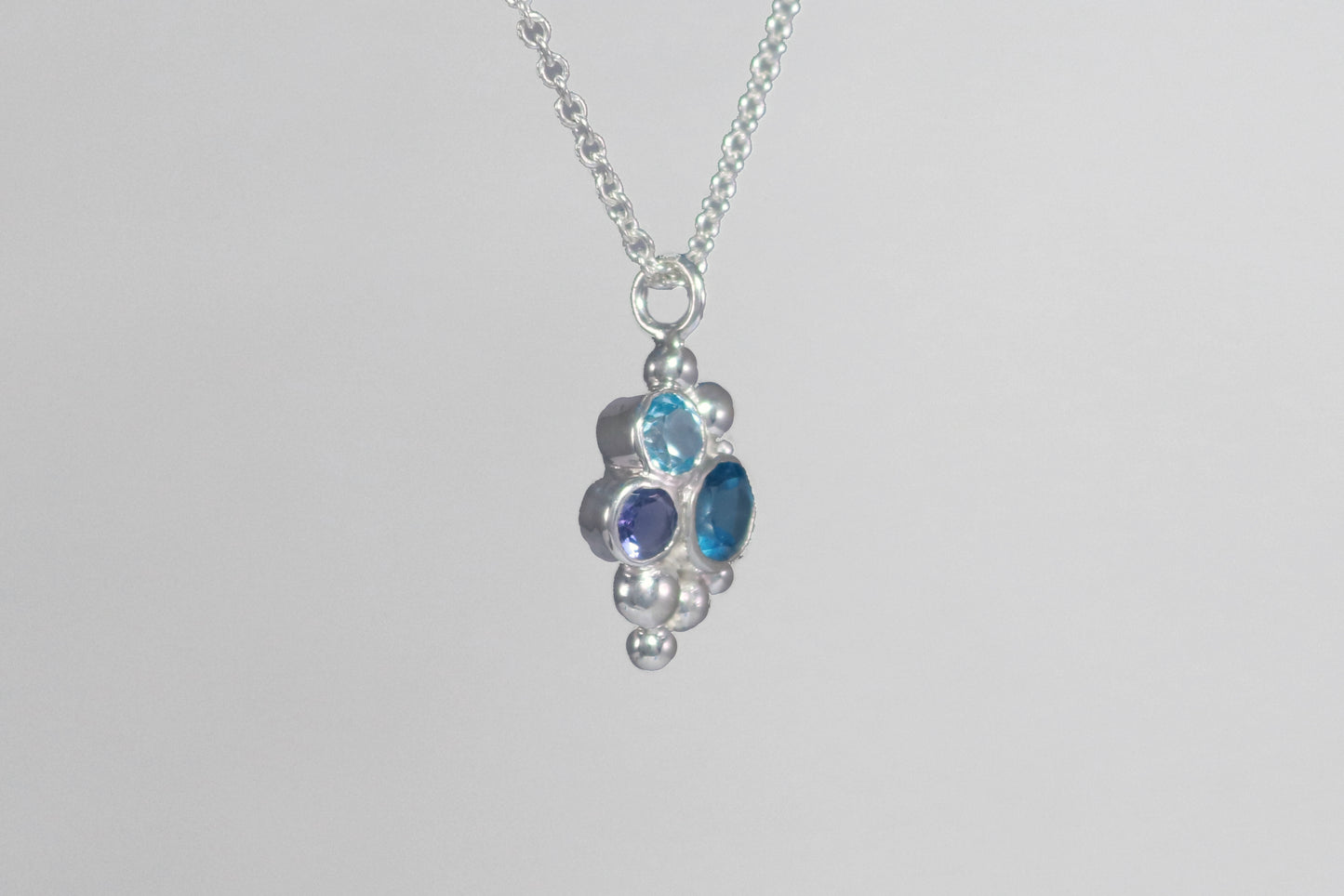 A pendant on a silver chain, three round gemstones, all a different shade of blue, set in a pyramid shape with randomly placed silver bubbles.