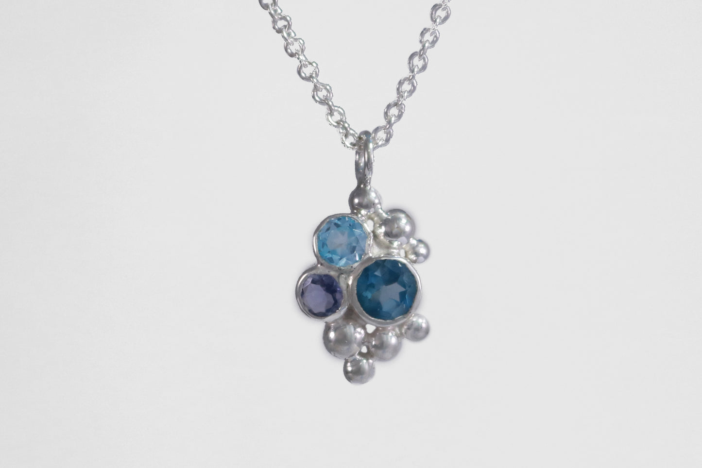 A pendant on a silver chain, three round gemstones, all a different shade of blue, set in a pyramid shape with randomly placed silver bubbles.