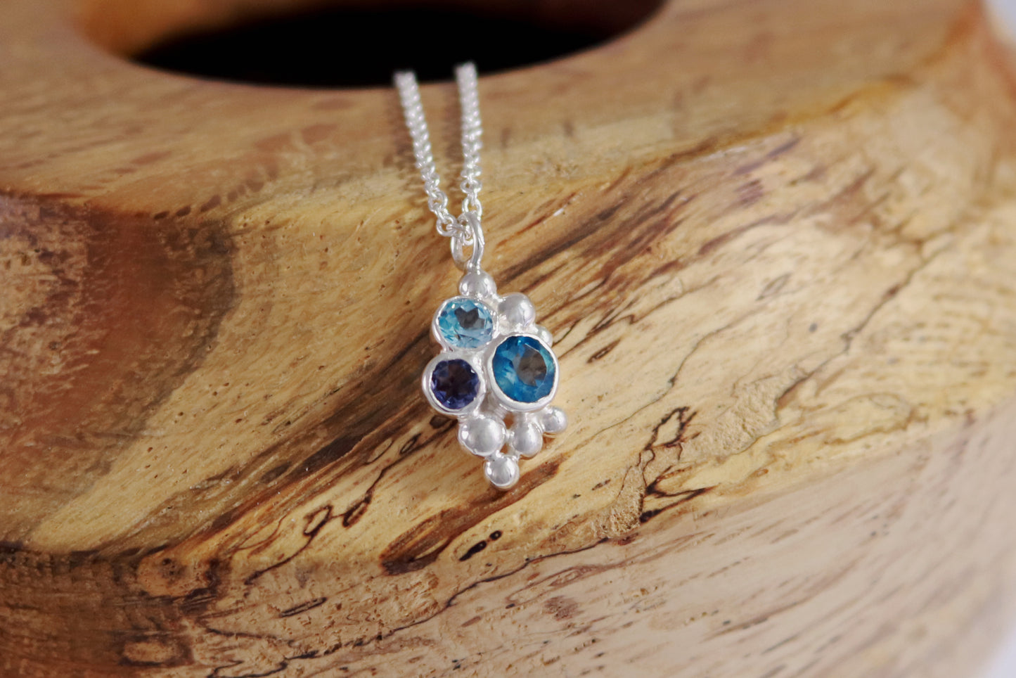 A pendant on a silver chain, three round gemstones, all a different shade of blue, set in a pyramid shape with randomly placed silver bubbles.