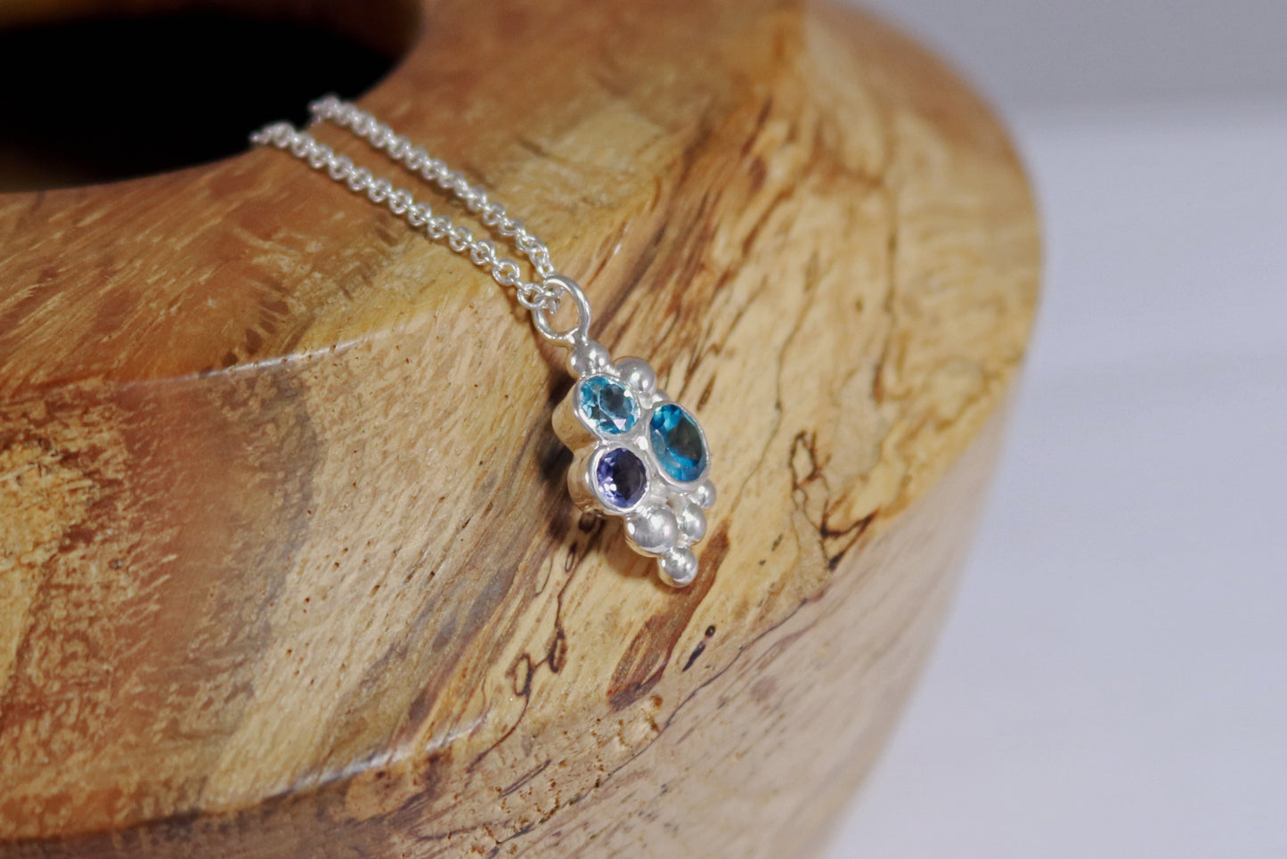 A pendant on a silver chain, three round gemstones, all a different shade of blue, set in a pyramid shape with randomly placed silver bubbles.