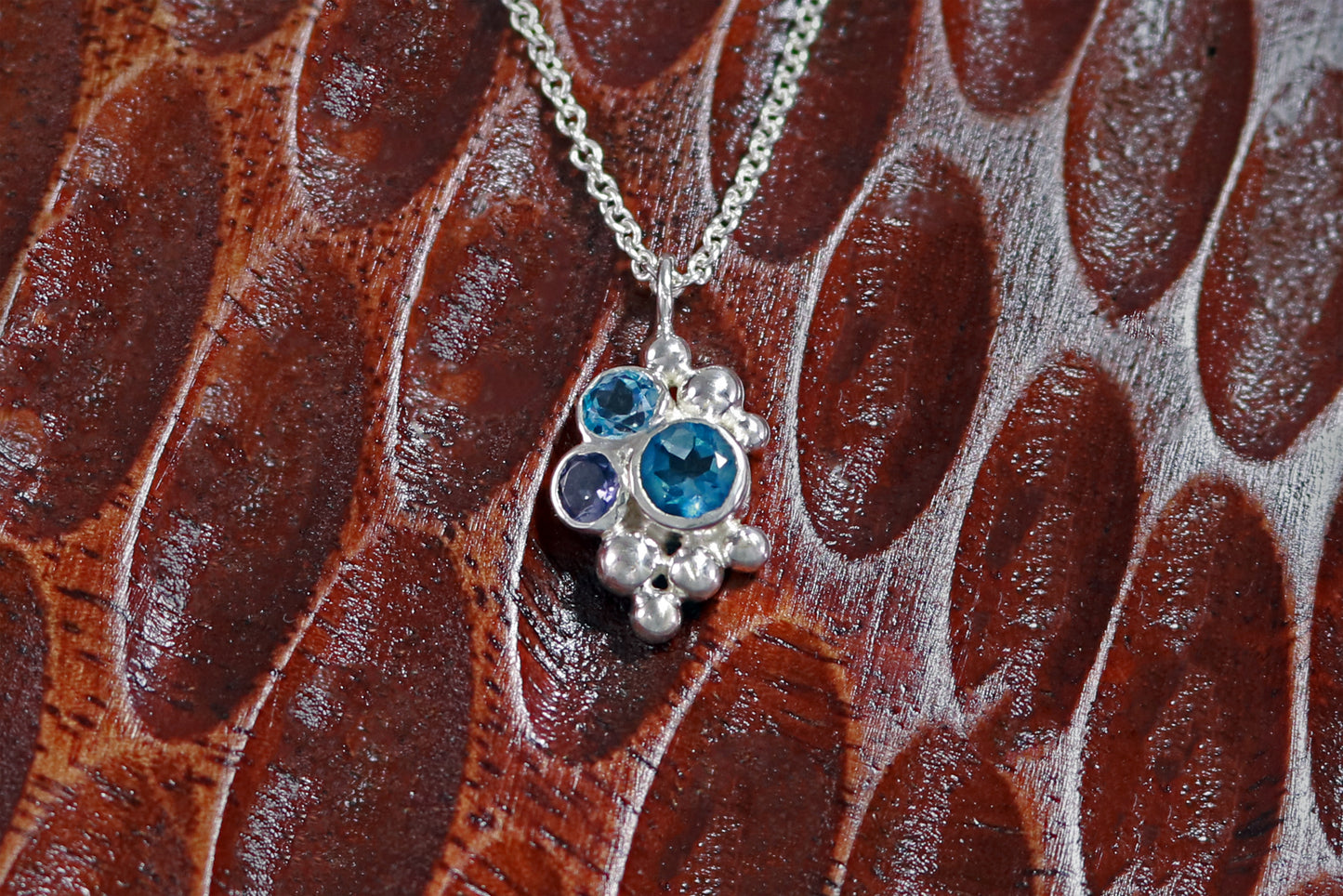A pendant on a silver chain, three round gemstones, all a different shade of blue, set in a pyramid shape with randomly placed silver bubbles.