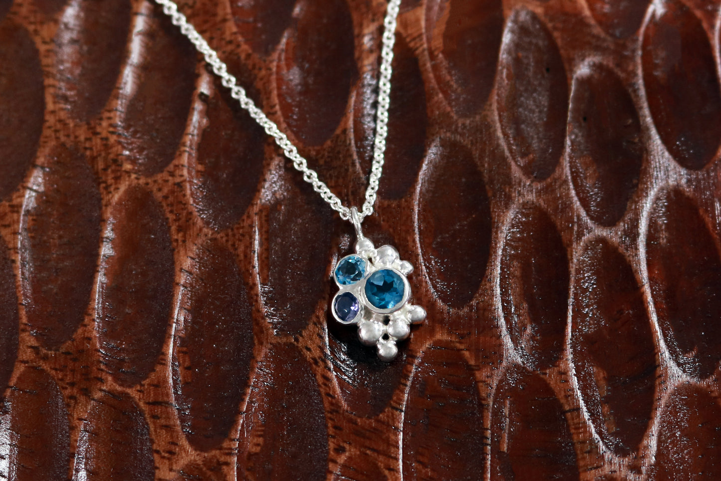 A pendant on a silver chain, three round gemstones, all a different shade of blue, set in a pyramid shape with randomly placed silver bubbles.