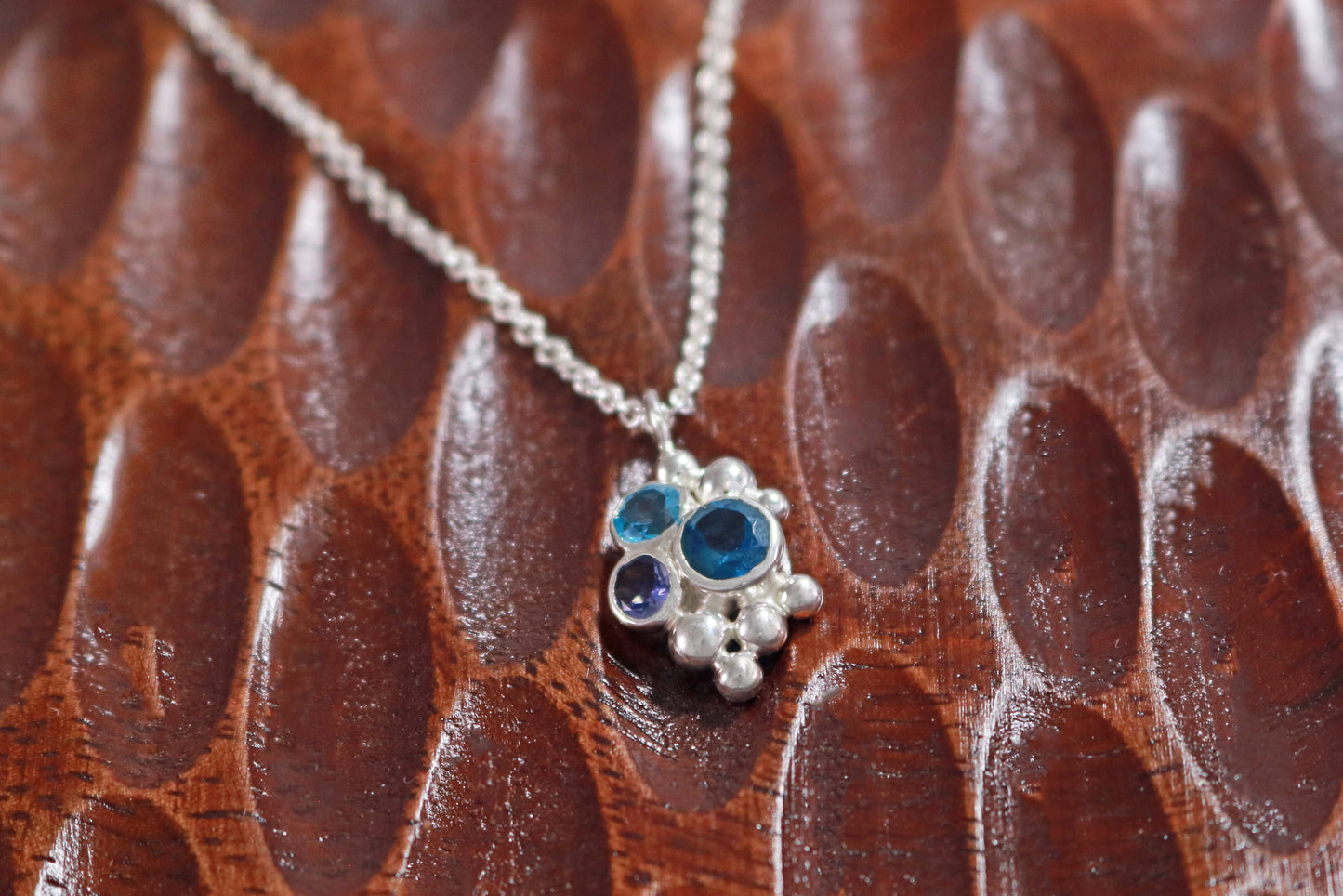 A pendant on a silver chain, three round gemstones, all a different shade of blue, set in a pyramid shape with randomly placed silver bubbles.