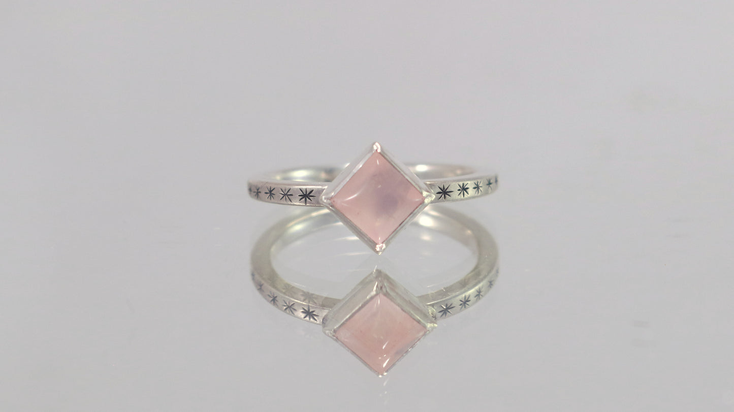 A sterling silver ring with a square rose quartz gemstone set in the center diagonally. Black stars going down each side of the ring band.