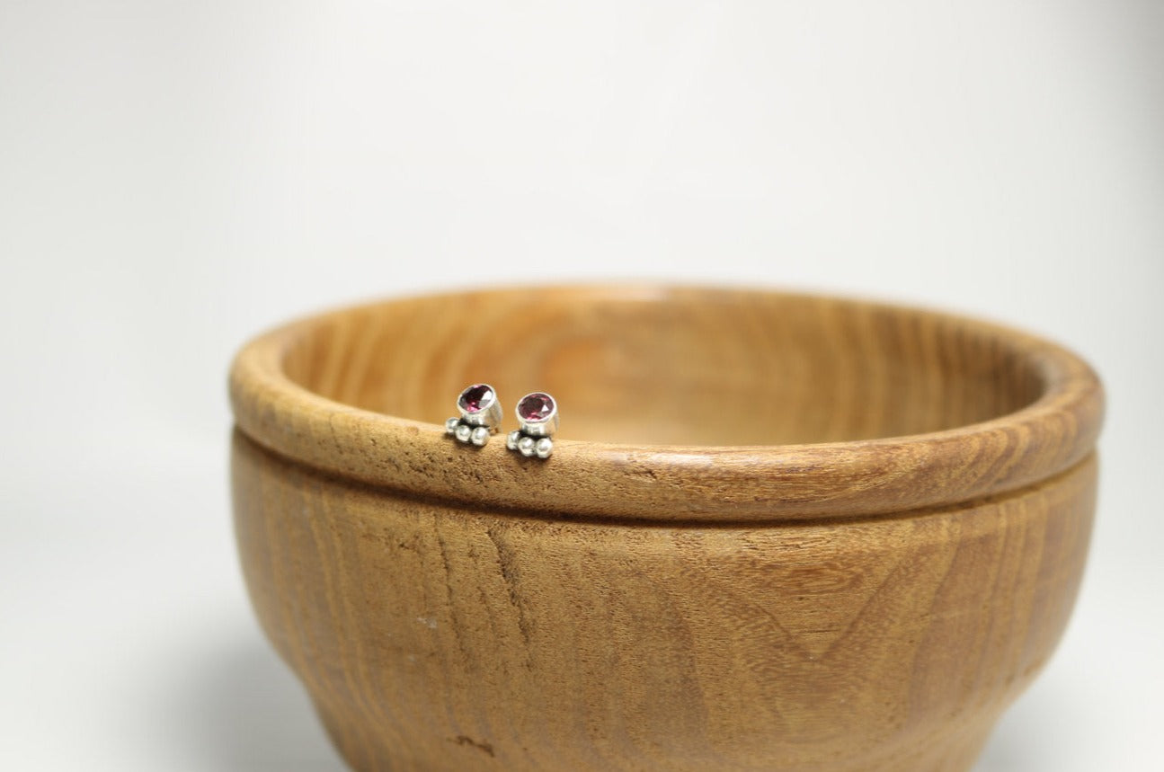 A pair of deep pink, round, faceted rhodolites set in sterling silver bezels with three silver bubbles aligned along the bezel.
