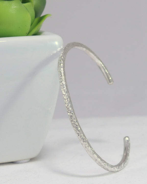 A solid sterling silver cuff covered in a small hammer texture, leaning against a succulent plant.