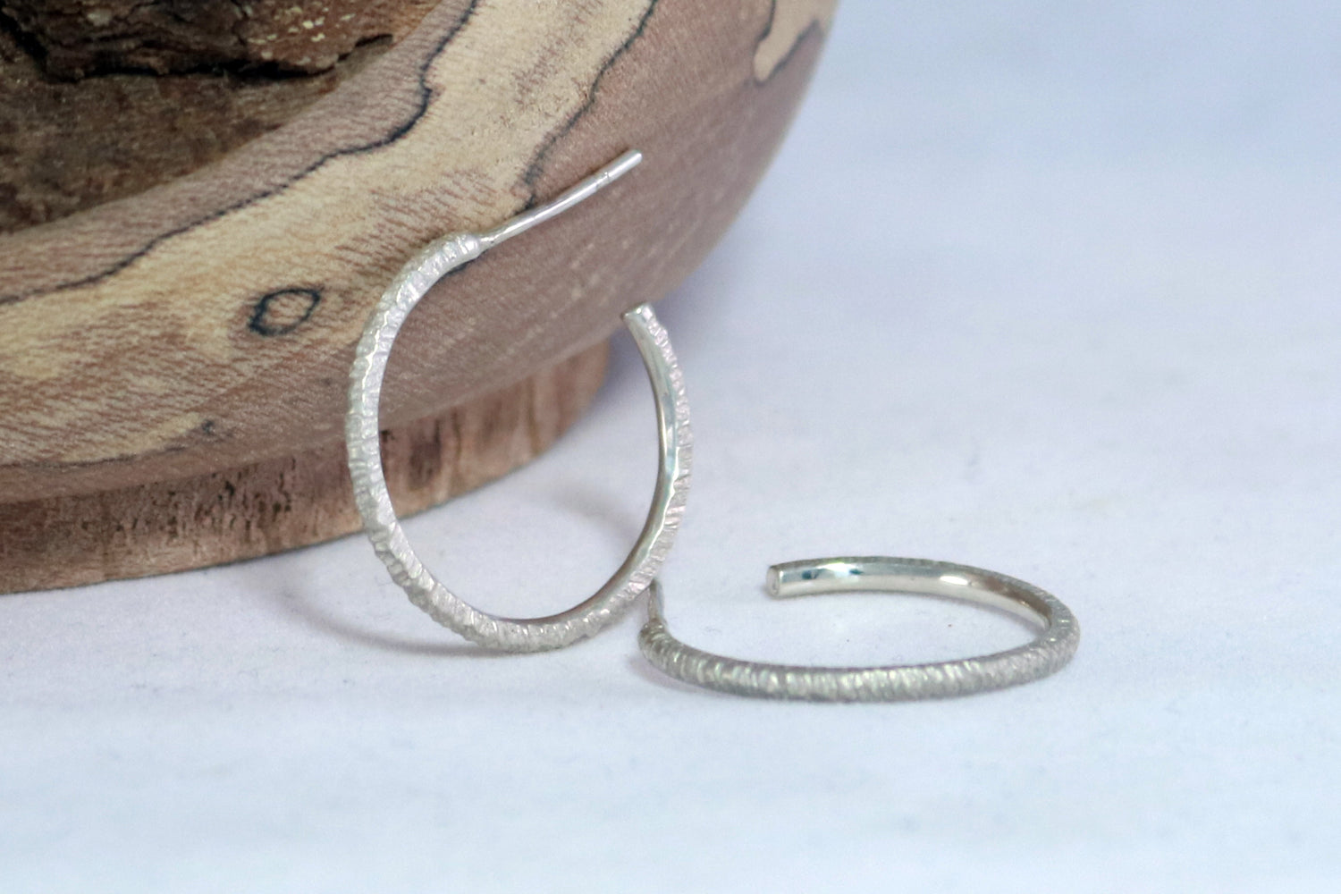 A pair of silver hoops leaning against a wooden bowl. The hoops are textured.