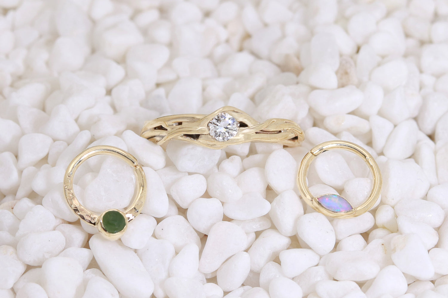 Three pieces sit on white pebbles. A jade and 14k yellow gold septum hoop, a 14k yellow gold and opal septum hoop and a 14k yellow gold and solitaire diamond one of a kind engagement ring featuring vines hugging a diamond.
