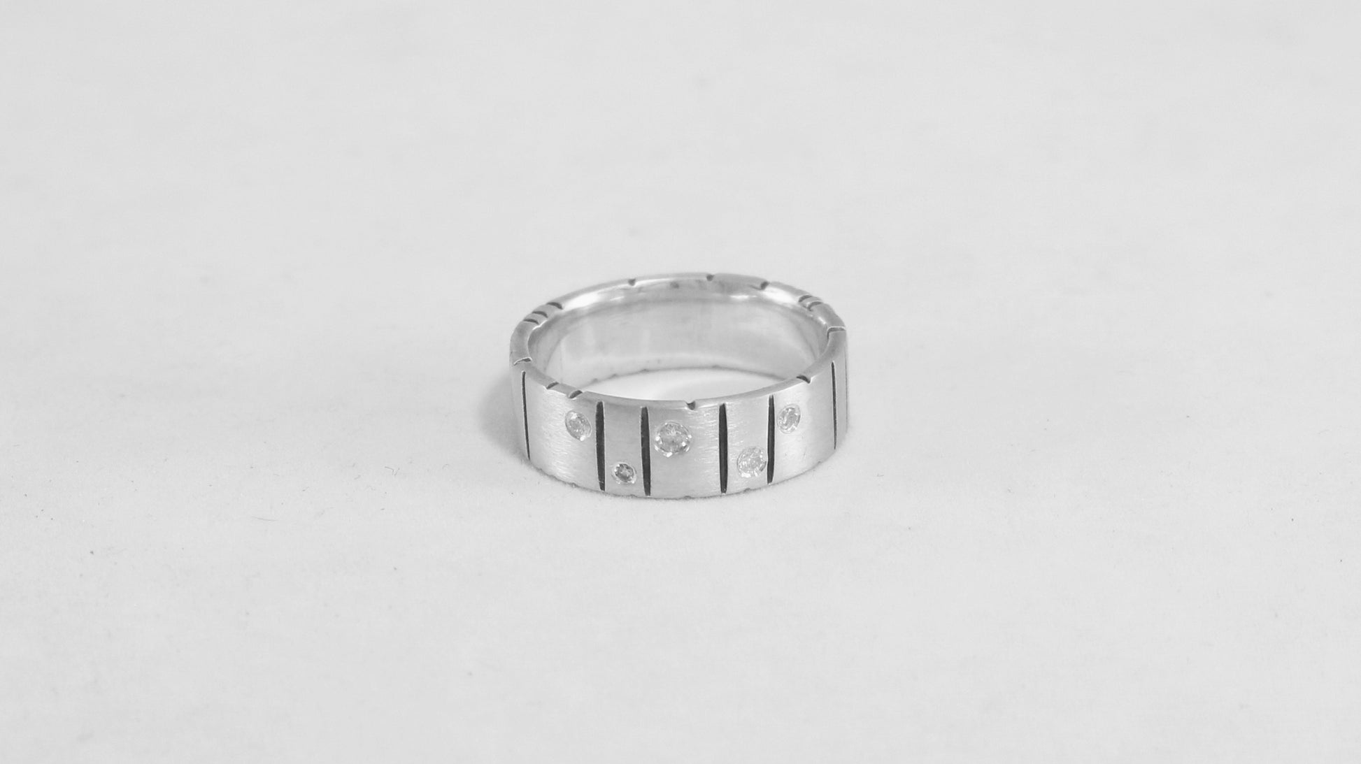 A thick silver ring band with randomly spaced black line details is flush set with 5 various sized diamonds.