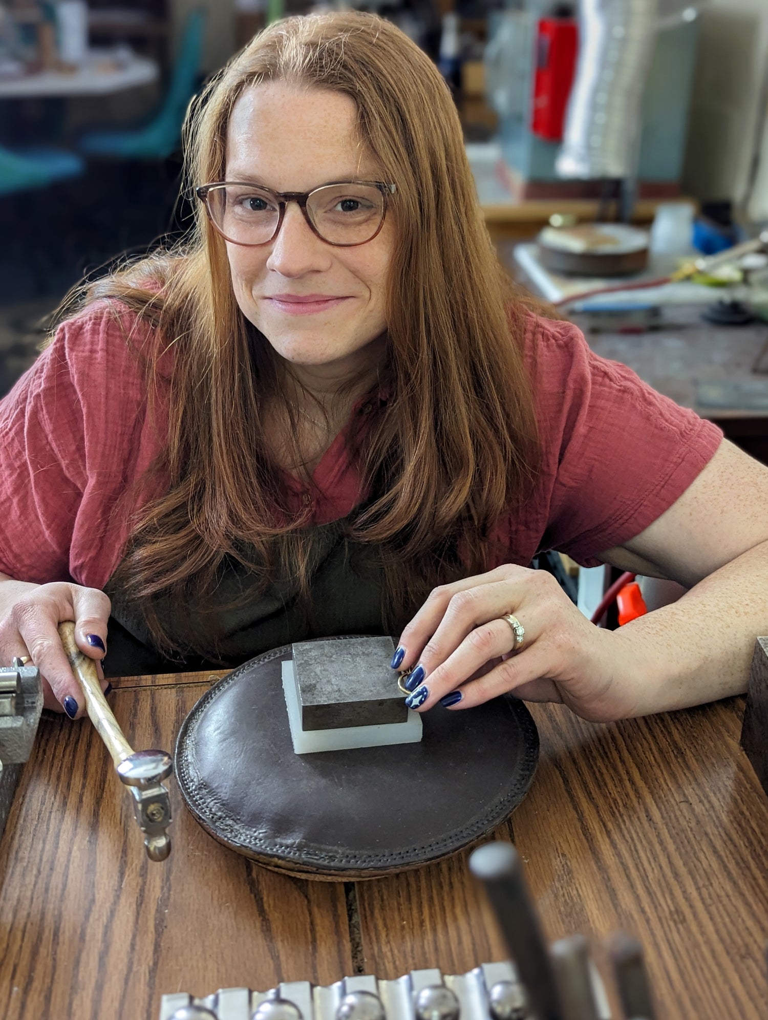 The goldmsmith, Tess Pell, is sitting at a table in front of her tools. She is holding a hammer and has long red hair, freckles and glasses. She is working on a ring and holding a hammer.