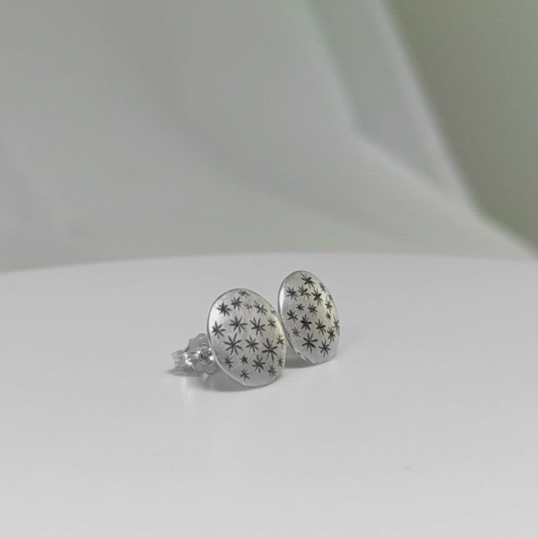 A video of a pair of round, domed, silver, stud earrings with hand carved black stars.