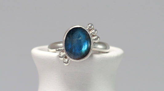 A royal blue, oval labradorite stone is bezel set on a sterling silver ring. On opposite diagonals are three silver bubbles with a total of 6 bubbles on the stone. 