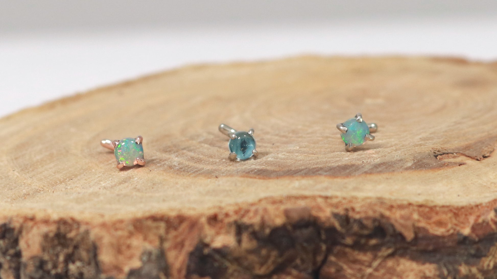 3 nose studs in a row on a piece of tree. From the left to right they are a rose gold nose stud with a round opal and 2 white gold nose studs, one with a round aquamarine and one with an opal, all 3 are set with prongs. 