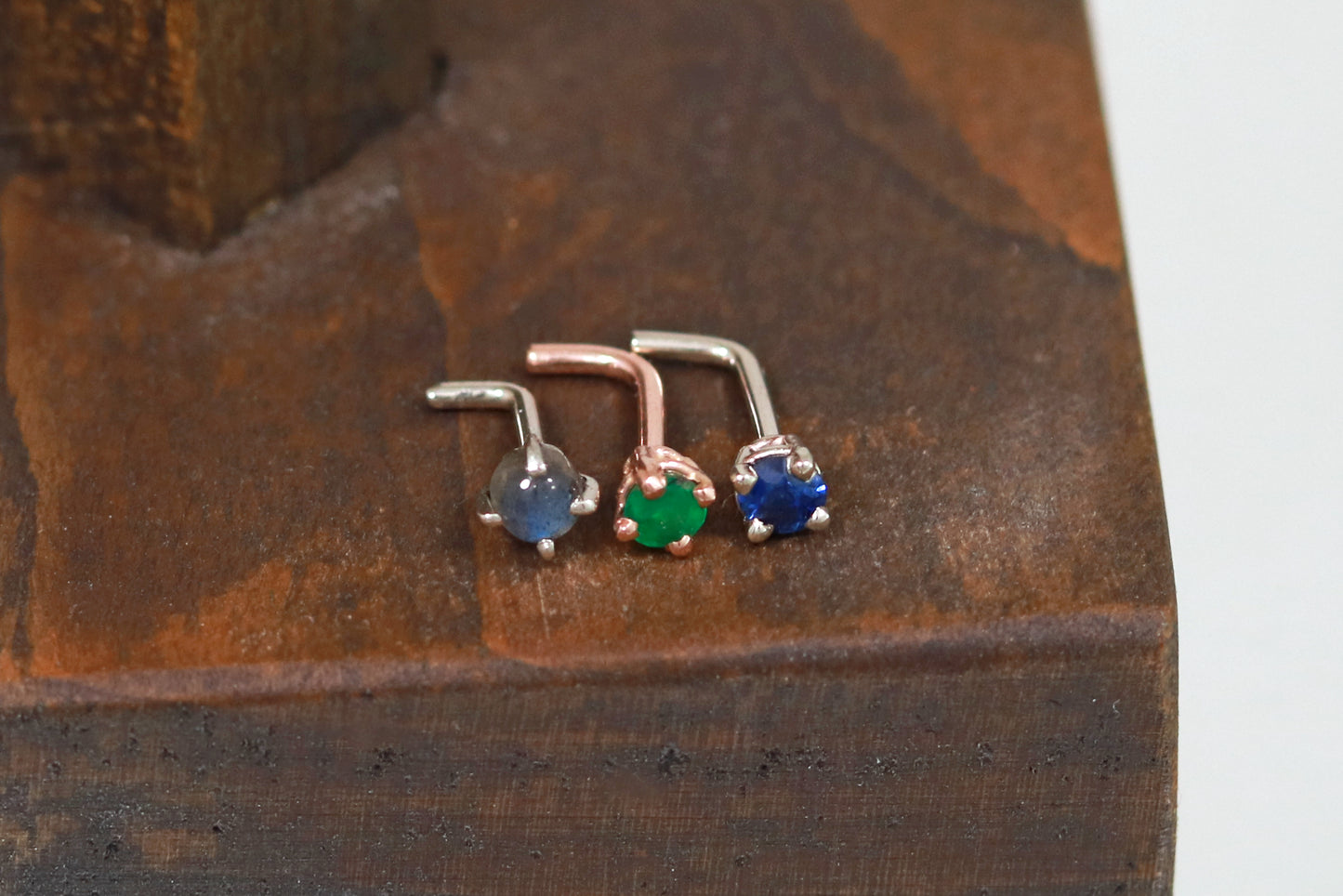 The top view of 3 nose studs in a row on a piece of dark wood. From the left to right they are a white gold nose stud with a prong set round rainbow moonstone, a rose gold nose stud with a basket set faceted emerald, and a white gold nose stud with a basket set faceted blue sapphire