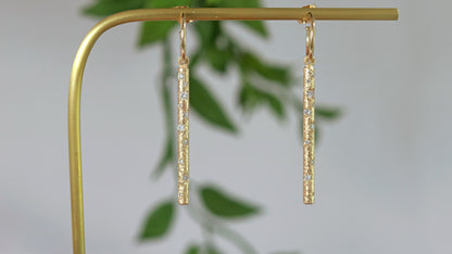 A pair of 14k yellow gold hoops with a solid yellow gold textured bar hanging from the hoop. Each bar has 10 diamonds of various sizes cascading down the bar.