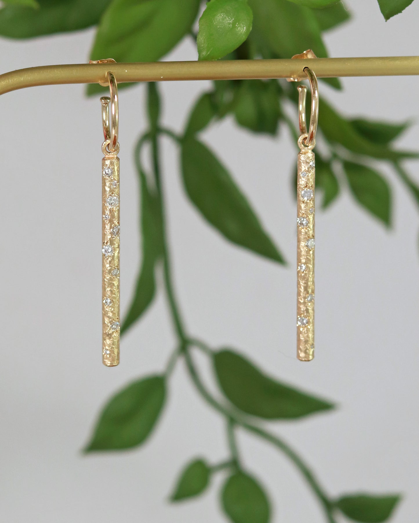 A pair of 14k yellow gold hoops with a solid yellow gold textured bar hanging from the hoop. Each bar has 10 diamonds of various sizes cascading down the bar. 