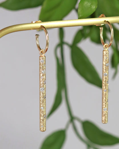 A pair of 14k yellow gold hoops with a solid yellow gold textured bar hanging from the hoop. Each bar has 10 diamonds of various sizes cascading down the bar.