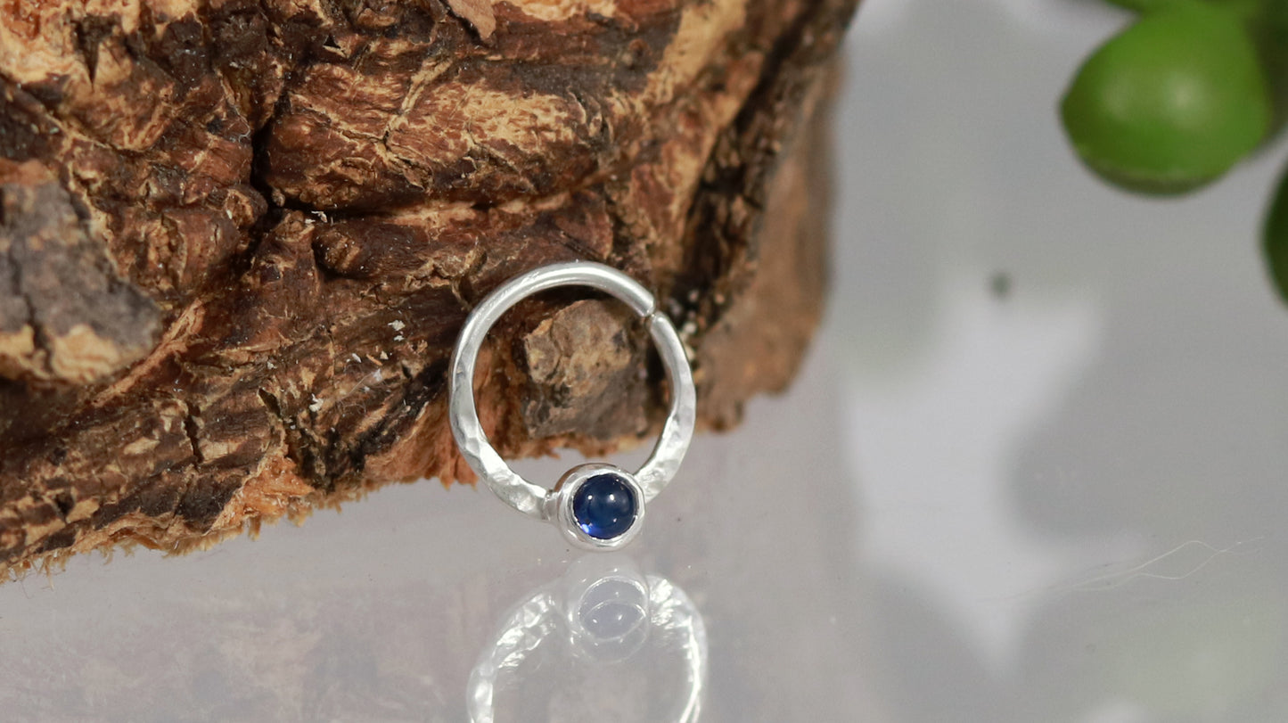 A sterling silver hoop, flattened and textured at the bottom, with a blue sapphire set in a bezel on top of the textured flat section.