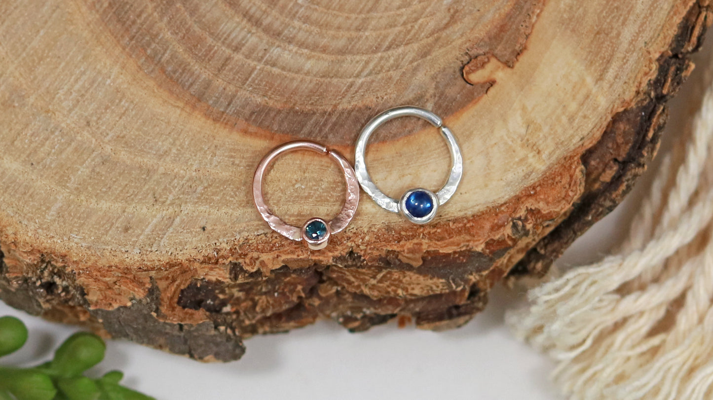 A sterling silver hoop, flattened and textured at the bottom, with a blue sapphire set in a bezel on top of the textured flat section. To the right of the silver hoop is an identical hoop made from solid 14k rose gold, set with a teal sapphire stone.