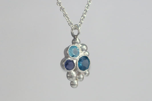 A pendant on a silver chain, three round gemstones, all a different shade of blue, set in a pyramid shape with randomly placed silver bubbles.