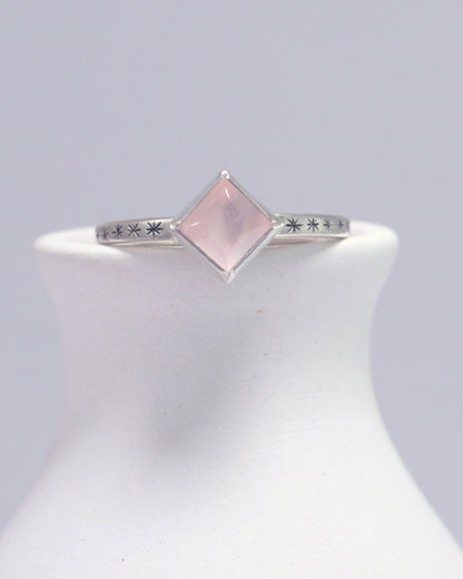 A sterling silver ring with a square rose quartz gemstone set in the center diagonally. Black stars going down each side of the ring band.