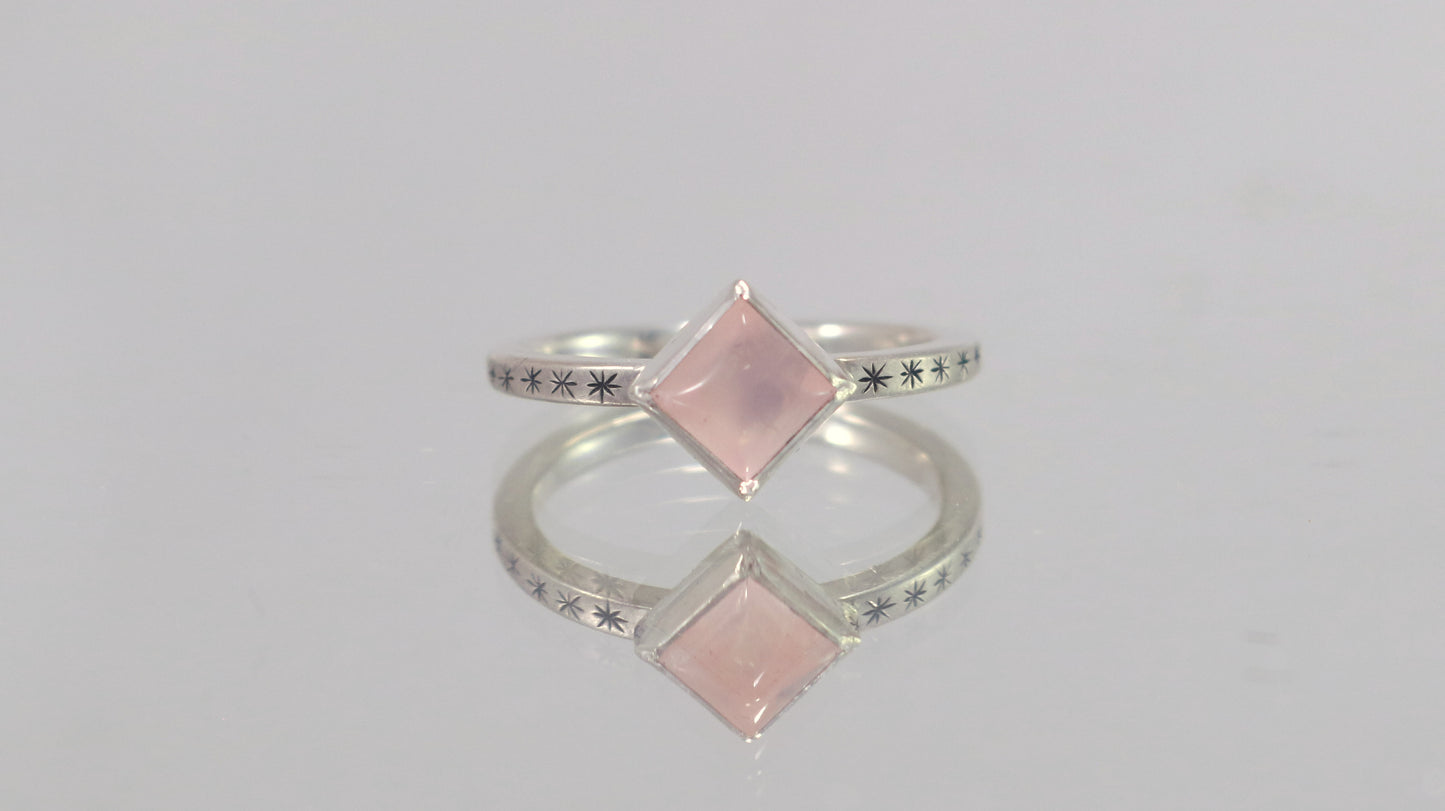 A sterling silver ring with a square rose quartz gemstone set in the center diagonally. Black stars going down each side of the ring band.