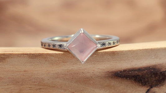 A sterling silver ring with a square rose quartz gemstone set in the center diagonally. Black stars going down each side of the ring band.