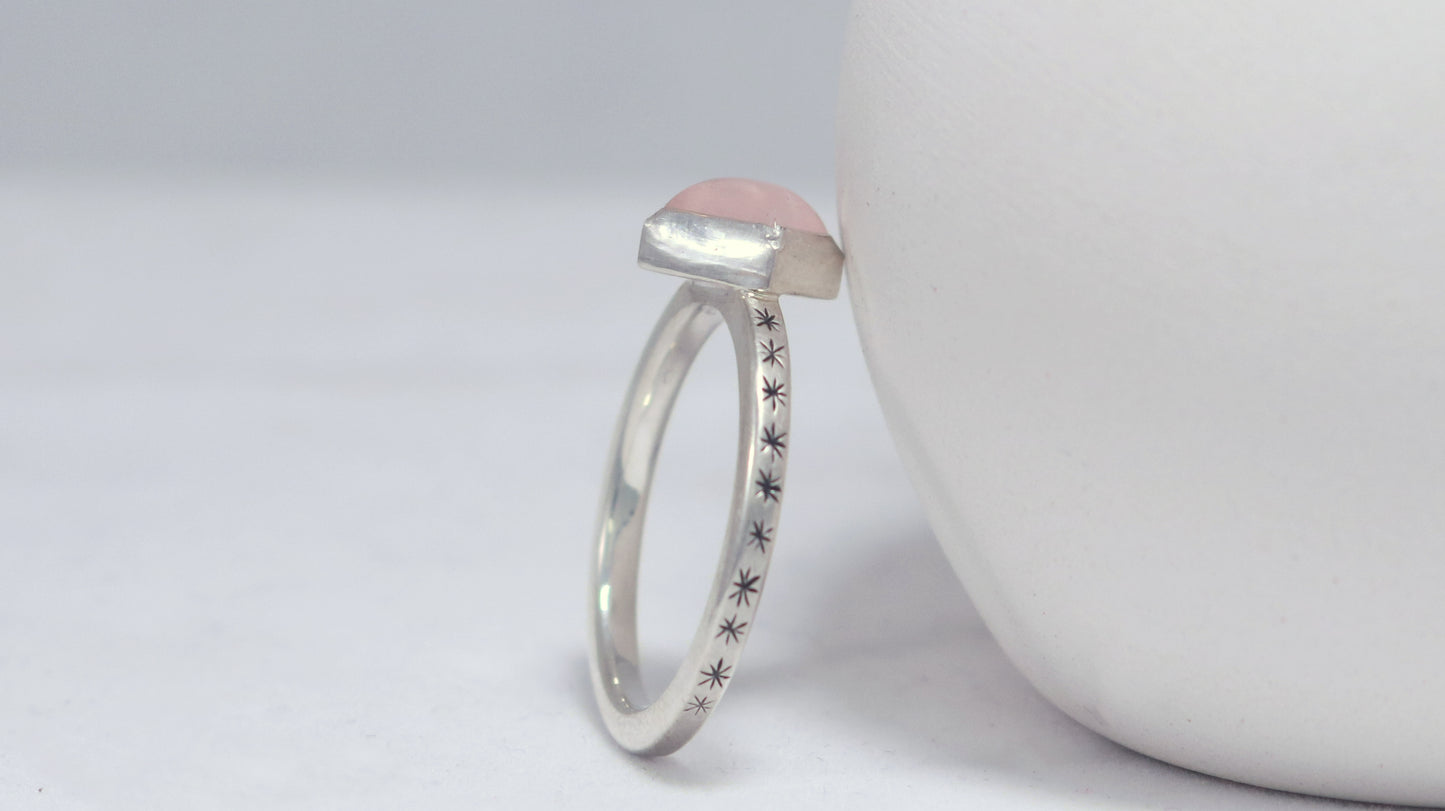 A sterling silver ring with a square rose quartz gemstone set in the center diagonally. Black stars going down each side of the ring band.