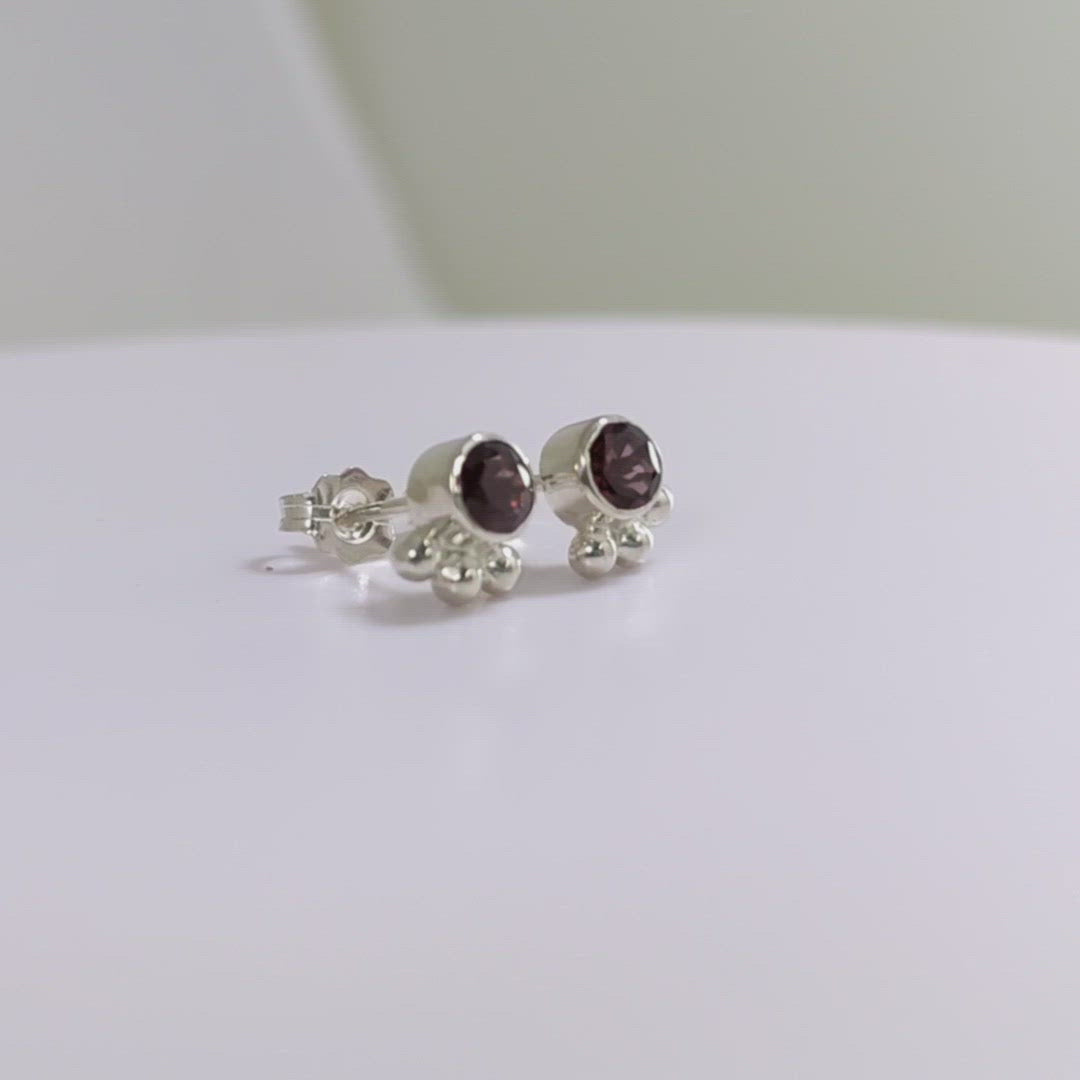 A video of a pair of deep pink, round, faceted rhodolites set in sterling silver bezels with three silver bubbles aligned along the bezel.