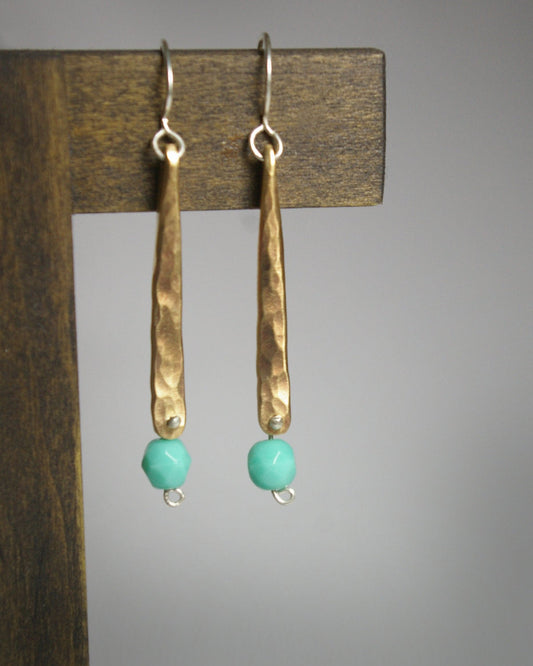A hand forged, textured, brass, elongated drop with a hanging glass turquoise colored bead.